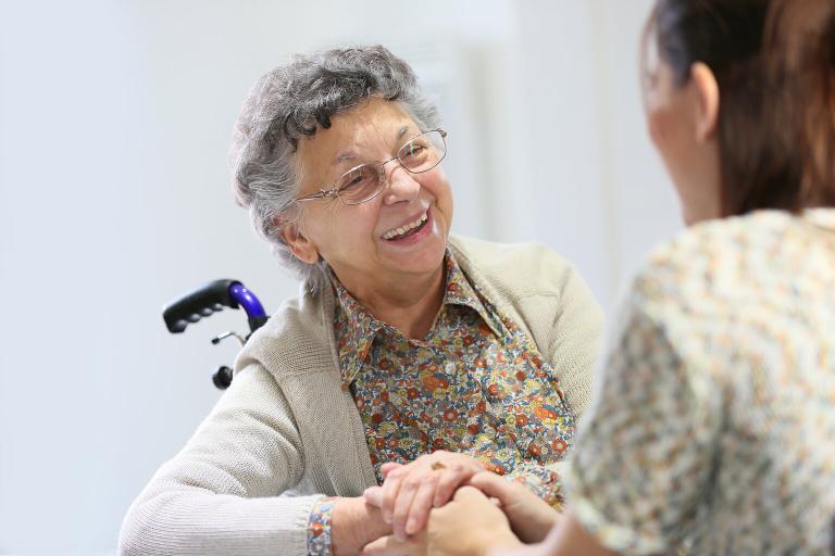  A photograph of an elderly lady in a wheelchair smiling at a carer who is holding her hand. The carer has her back to the camera.