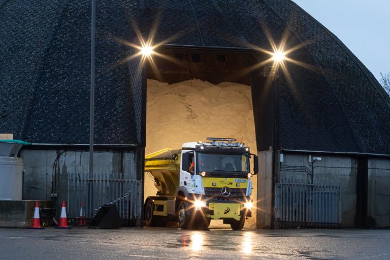 A gritting lorry with its lights on, driving out of a large domed salt barn structure.