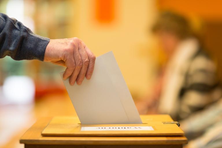 A close up of a man's arm depositing a piece of folded paper in a ballot box