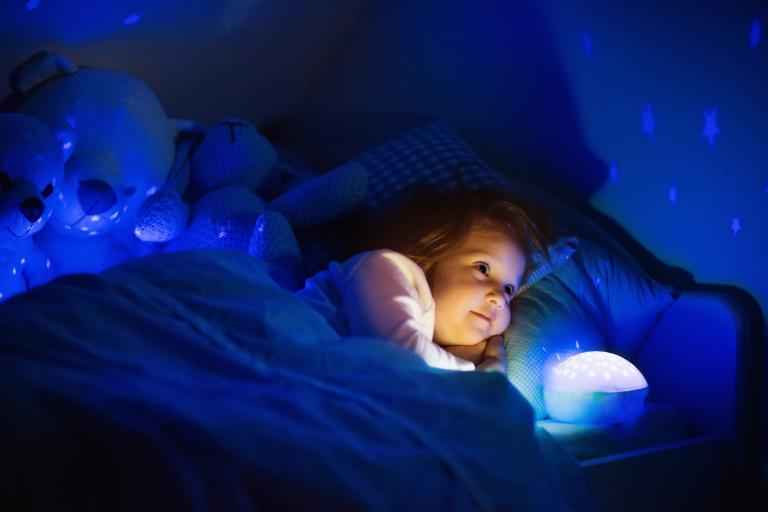 Little girl laying in bed with a night light projecting stars on the ceiling