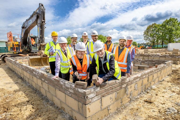 A group of people wearing yellow and orange high visibility jackets and hard hats standing behind a brick wall, which is being constructed on a building site.