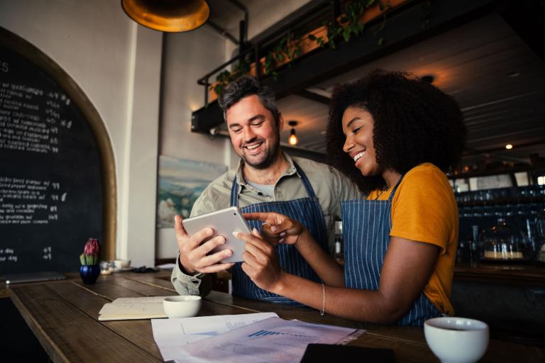 A man and woman behind the counter of a coffee shop looking at a tablet.