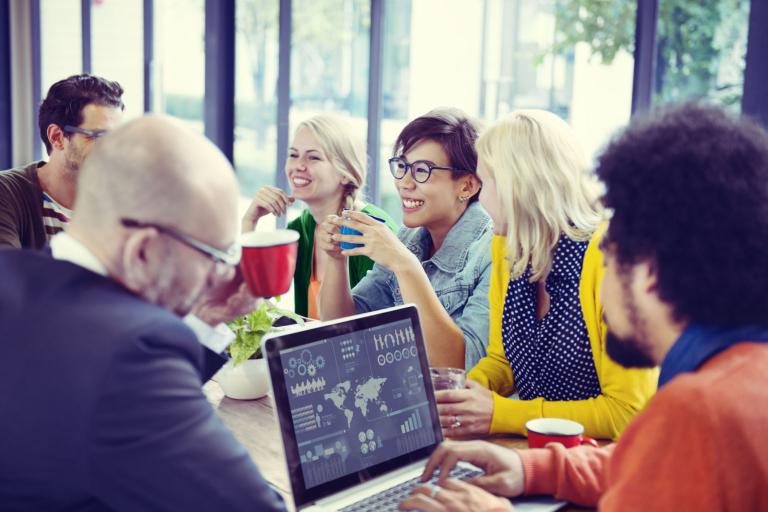 A group of people sitting around a table in discussion, with coffee mugs and a laptop on the table and in their hands.