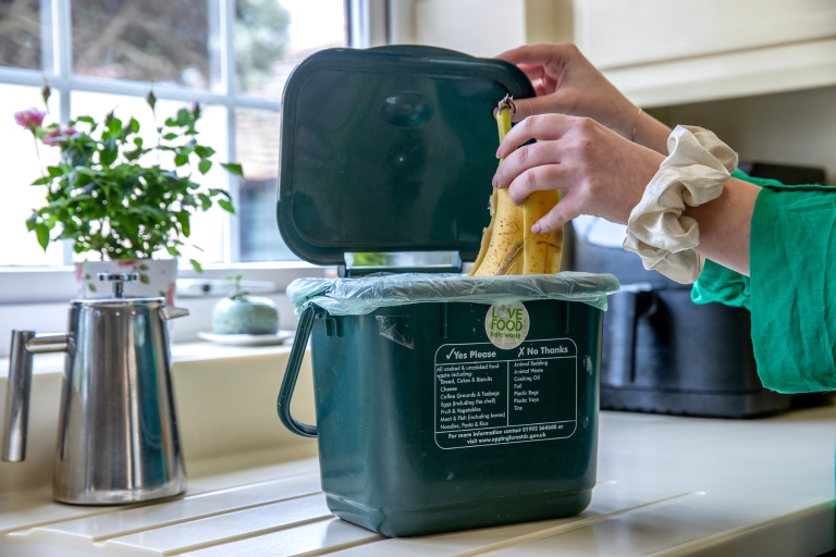 A hand placing a banana peel into a food recycling caddy on a kitchen countertop in front of a plant and a window.