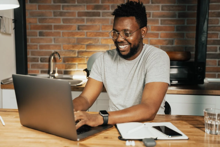 A smiling young man with glasses, typing on a laptop. A notebook, mobile phone and a glass of water are on the table next to him.