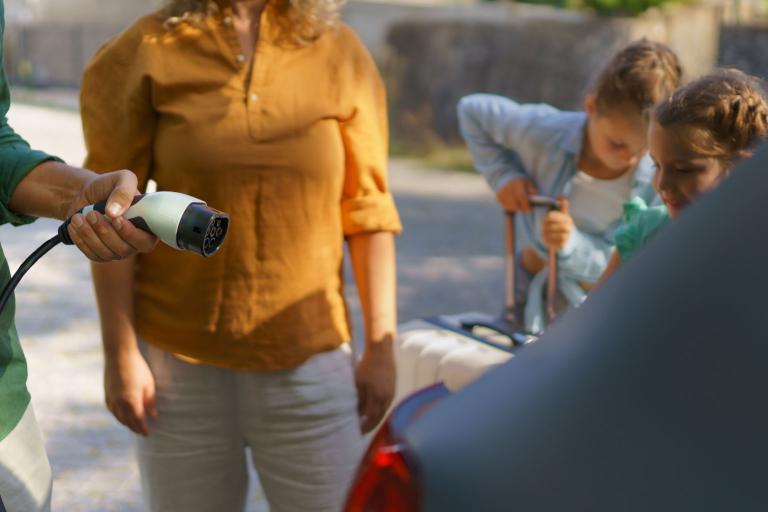 A family gathered by the boot of a car. Two children, one with a suitcase, a mother and a father holding an electric vehicle charger. 