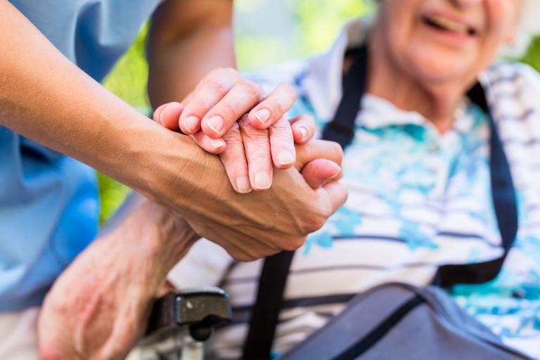 A carer in a medical gown holding the hand of an elderly person, who is sitting in a wheelchair.