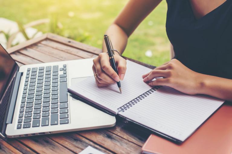 A woman sitting outside on a wooden table writing in a lined notebook, which is resting on an Apple Macbook computer.