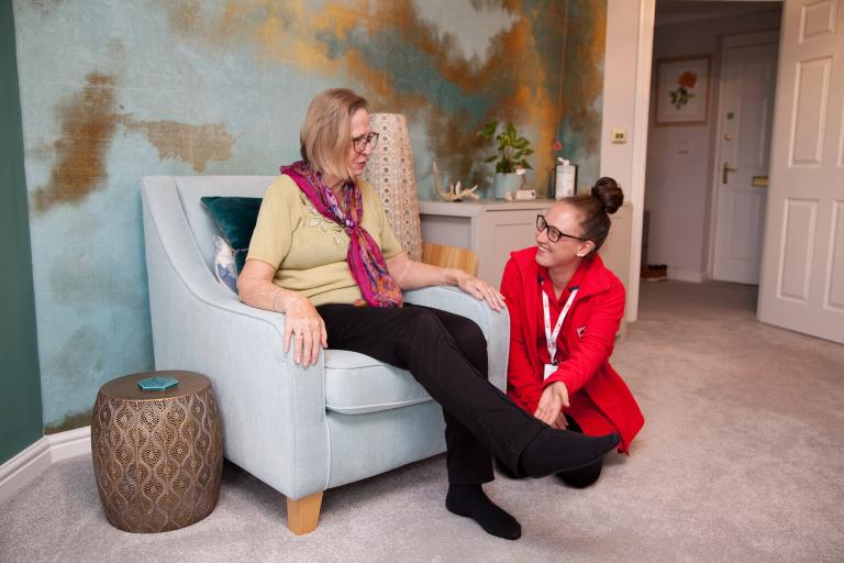 A lady is sitting in a blue armchair in a living room. A care worker wearing red is kneeling next to her and holding her foot. 
