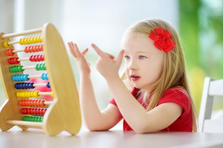 A young girl with blonde hair and eyes is counting on her fingers. She has an abacus in front of her. She is sitting at a desk and she has a large red bow in her hair.