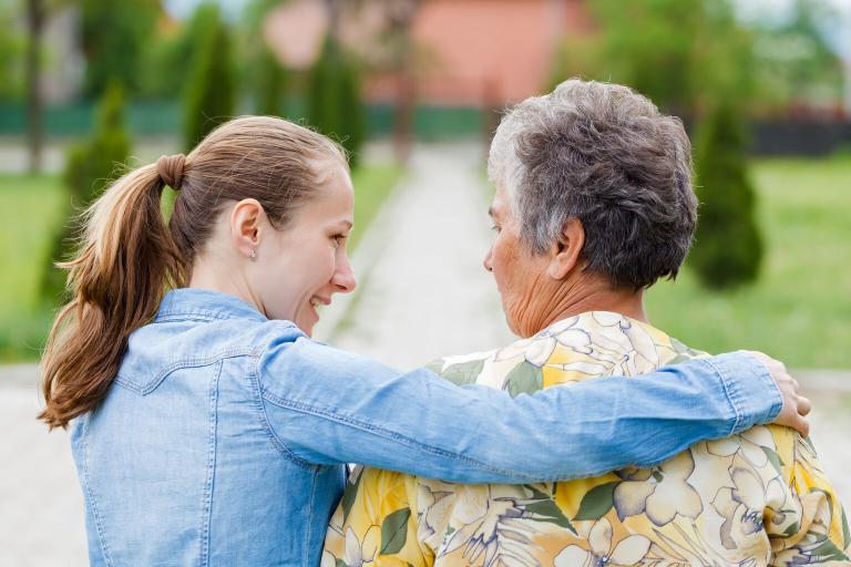 A photograph of a young carer with her arm round an old lady. Their backs are to the camera and they are walking along a path in park.