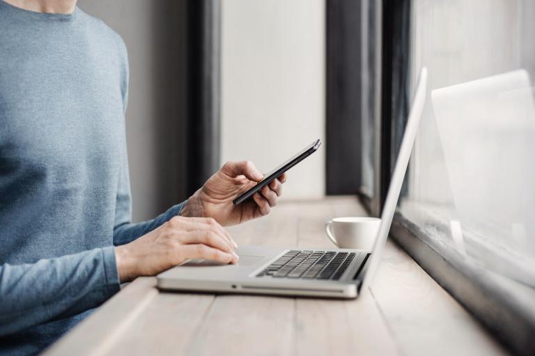 A man on a laptop typing with one hand and his phone in the other hand.