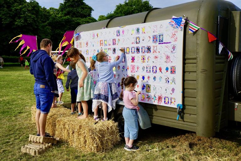 A photograph of five children standing on hay bales drawing on a large green box. This is at a festival with bright coloured banners and decorations. A man is standing and talking with the children.