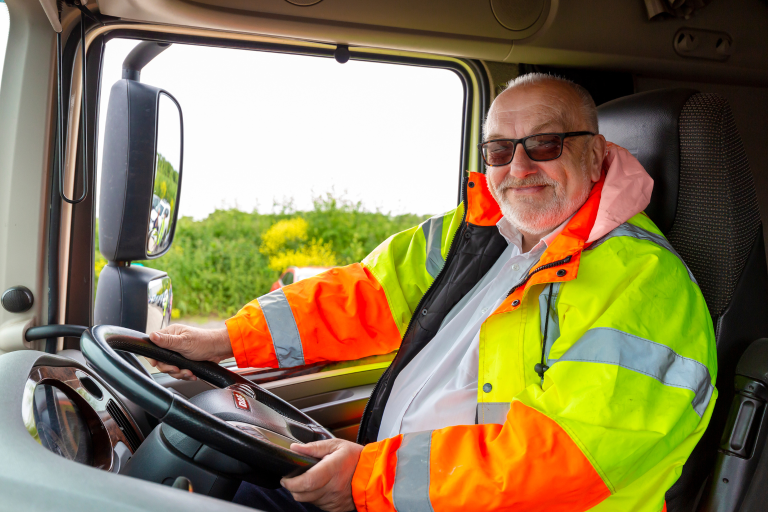 Councillor Tony Ball, Essex County Council Cabinet Member for Education Excellence, Lifelong Learning and Employability, smiling, wearing sunglasses and sitting behind the wheel of an HGV, wearing a yellow and orange high-visibility jacket. 