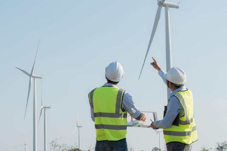 Two male engineers in yellow high-visibility jackets and white hardhats looking at a set of wind farm machinery. 