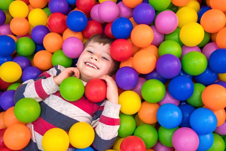 Little boy wearing striped top lies in colourful ball pool smiling.