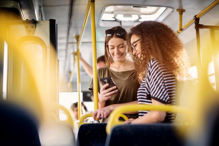 Two women aboard a bus. One is looking at her phone whilst holding on to a yellow handle on top of a seat. Both are smiling.