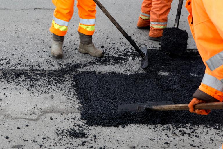 A close up of three workers in orange high-visibility clothing spreading asphalt on a road. 