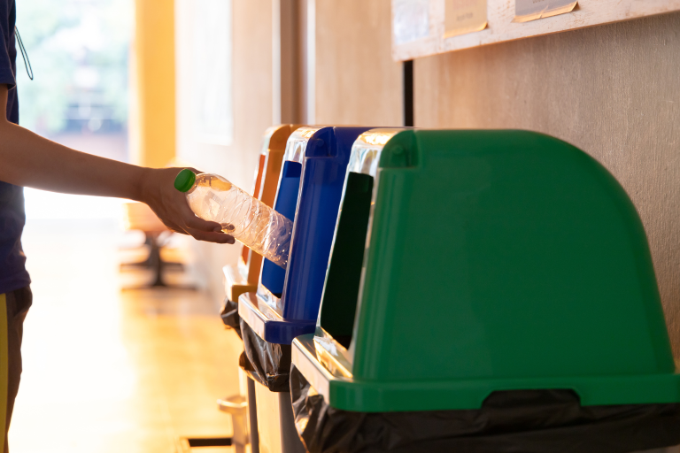 A person depositing a plastic bottle in a blue recycling bin, which is in between green and yellow recycling bins. 