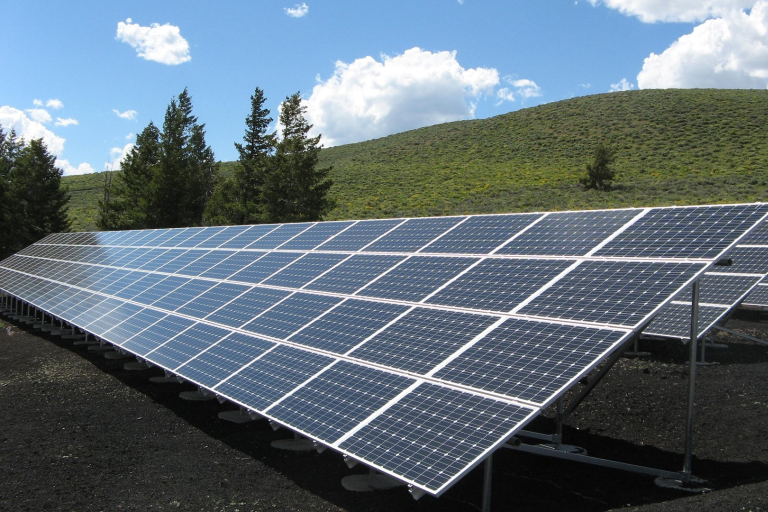 A large collection of solar panels on a green hillside featuring a number of trees, with a blue sky and clouds behind. 