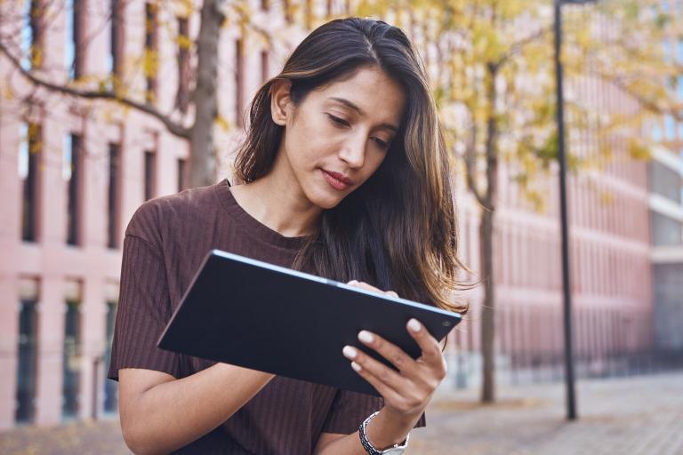 A lady with long dark hair wearing a brown t shirt is sitting on a step outside a building. She is looking down at her tablet and is typing.