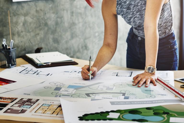 A woman with pieces of paper, blueprints and plans on a desk in front of her. She is holding a pen.