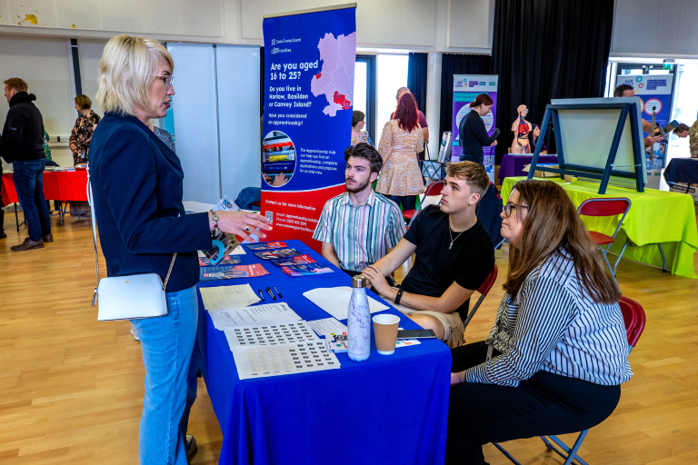 Three people sat behind stall at a careers fair talking to blonde woman