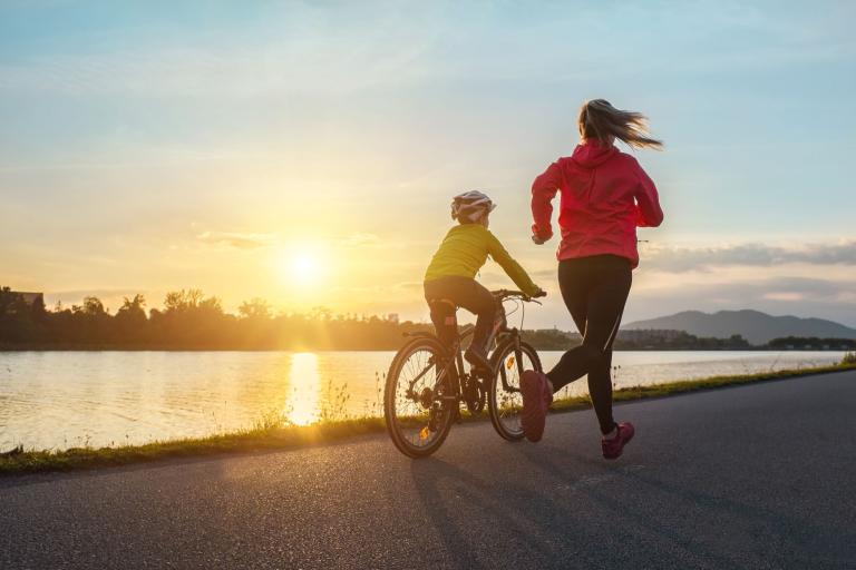 A boy wearing a helmet cycling alongside a woman, who is running next to him. In the background is a lake 