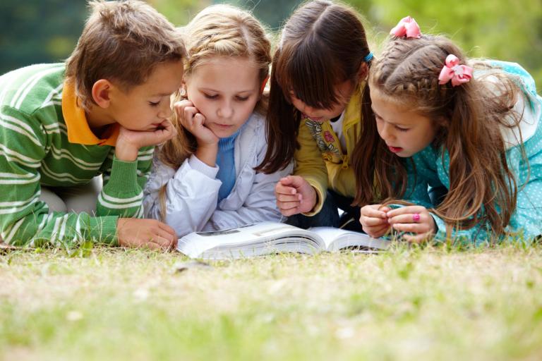 A boy and three girls reading a book outside on an area of grass.
