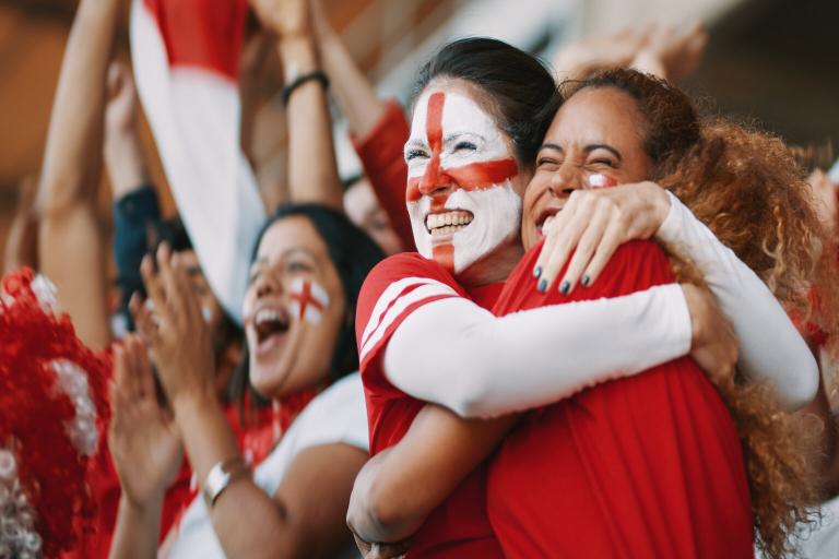 Two women hugging at a football match. They are England supporters and have the England flag painted on their faces.