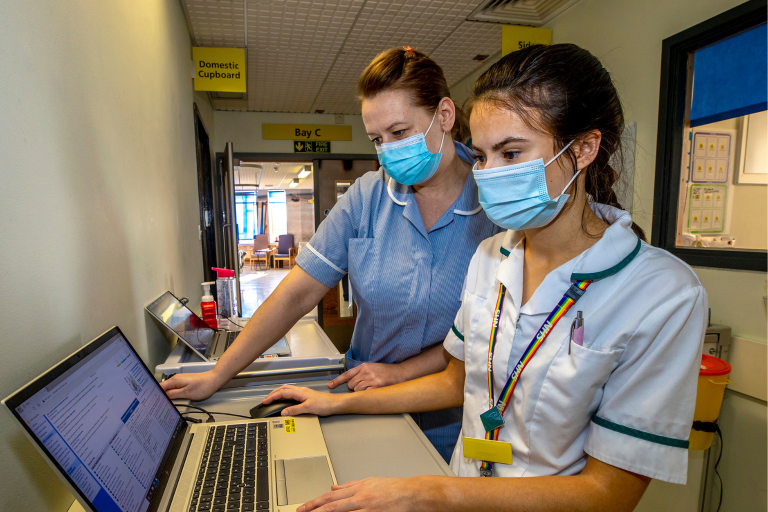 Two female members of healthcare staff wearing facemasks and looking at a laptop.