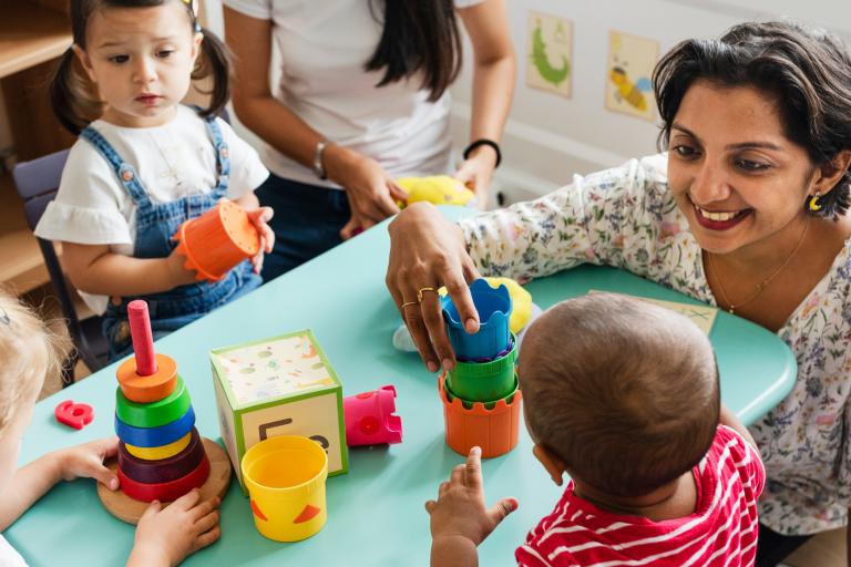 Early years practitioner sat with three children playing with toys on the table.