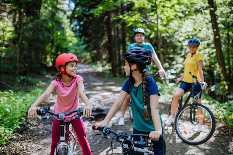 A mum and dad with two young girls in a woodland area, all wearing bicycle helmets and pushing their bikes.