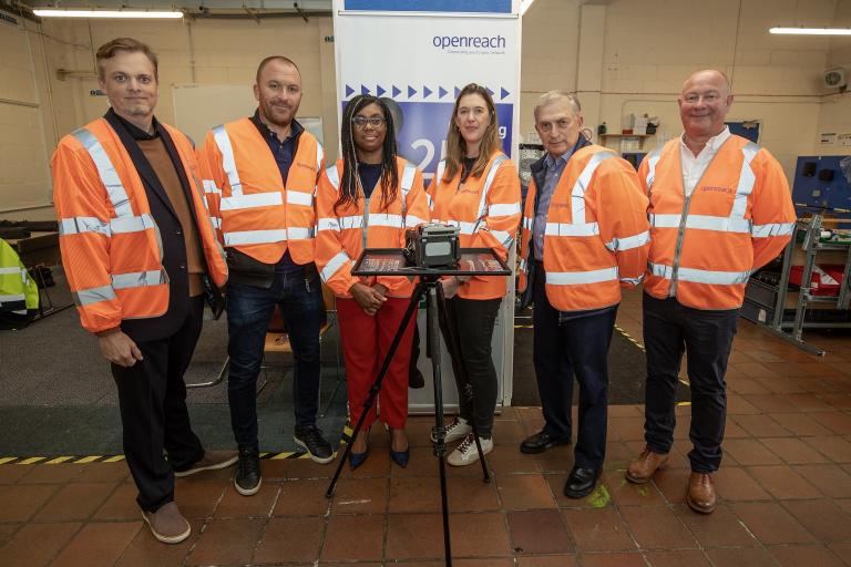 Six people wearing orange high-visibility jackets standing in front of a tripod with a piece of technical equipment on it.