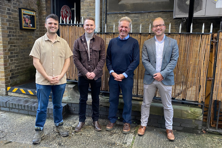 Four men stand in front of a brick wall while smiling to camera.