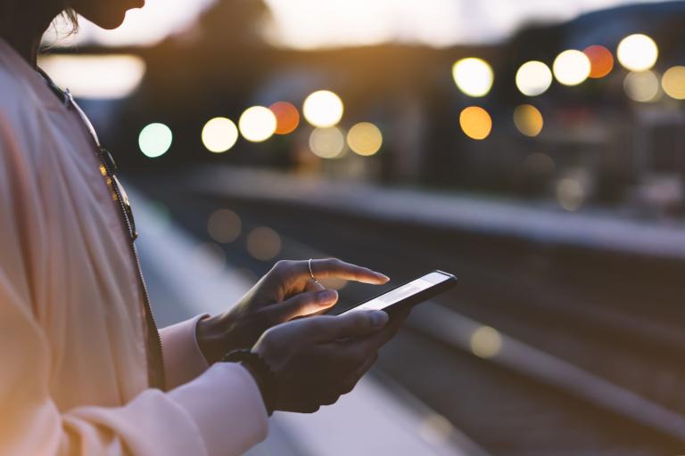 A woman standing on a station platform in the evening, holding her smartphone.