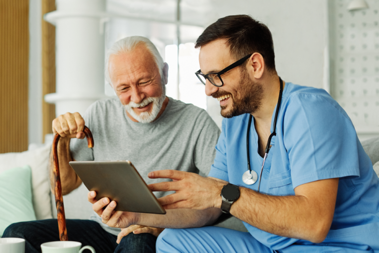 Senior man and medical professional sit on a sofa while smiling and view electronic tablet together.
