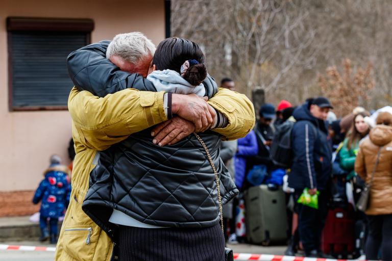 A man and woman dressed in winter clothes embrace in the foreground as refugees queue with luggage in the background.