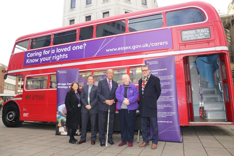 A red old double decker bus. It has branding on teh side that reads Caring for a loved one? Know your rights. It also has the website address www.essex.gov.uk/carers. A group of people are standing in front of the bus smiling. The bus is at the front of County Hall, a large building with big steps to the entrance.