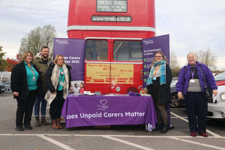 A red double decker bus branded with teh text 'Care for a loved one? Know your Rights'. There is a group of people standing in front of the bus with an information table,