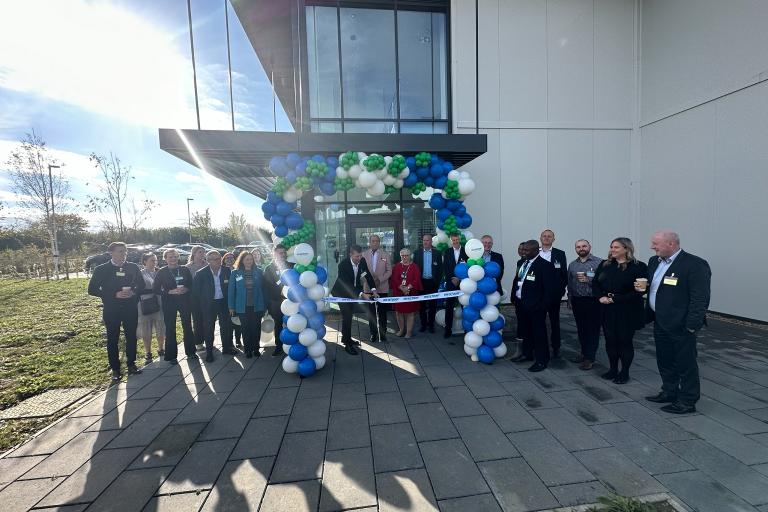 A large group of people gathered around a balloon archway, with a ribbon being cut. 