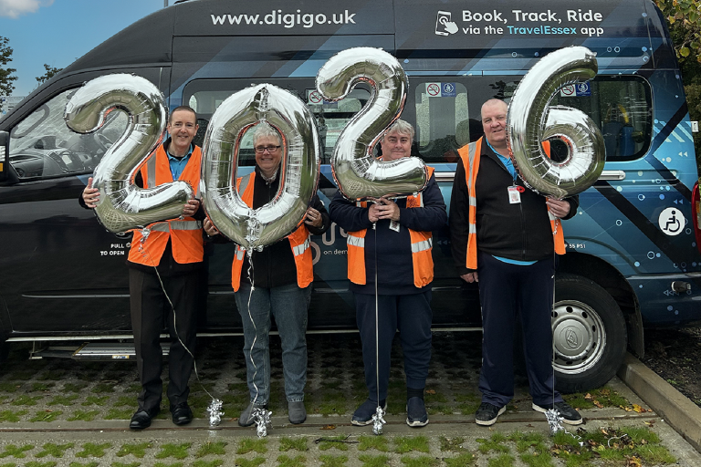 Four people standing in front of a DigiGo minibus wearing orange high-visibility jackets and holding balloons spelling out 2026.