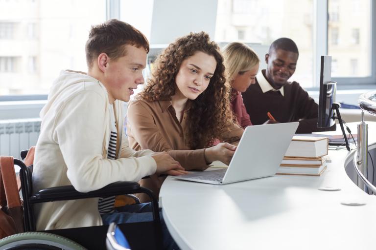 Young man in a wheelchair views a laptop with assistance from a young woman in an office setting.