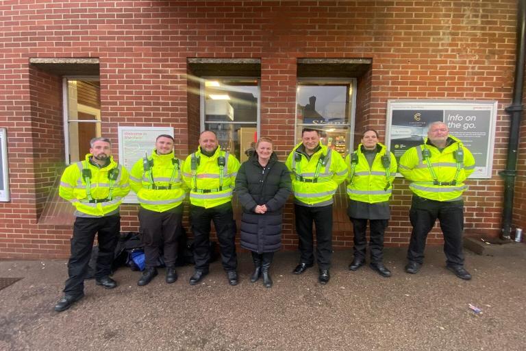 Cllr mckinlay standing with six new officers who are wearing uniform and yellow coats. They are standing in front of a brick wall and are all smiling.