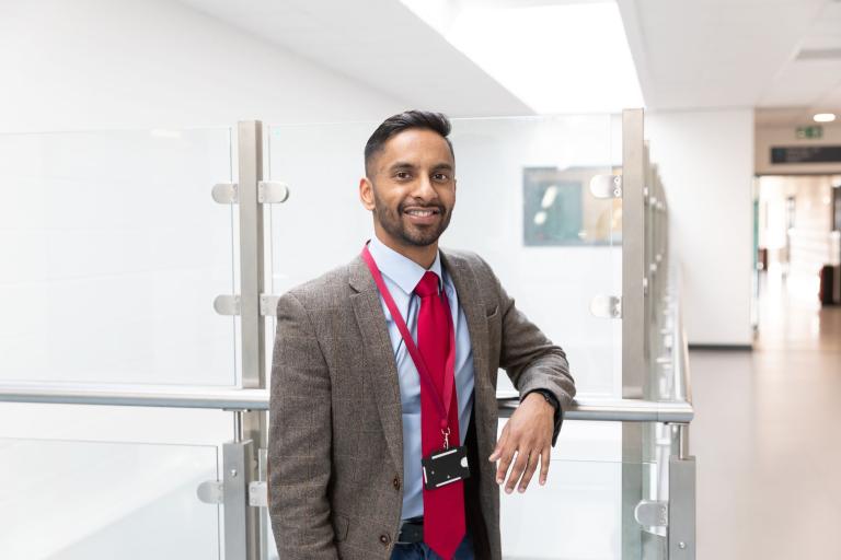 Bobby Seagull stood in a school corridor, he's wearing a red tie and a brown jacket.