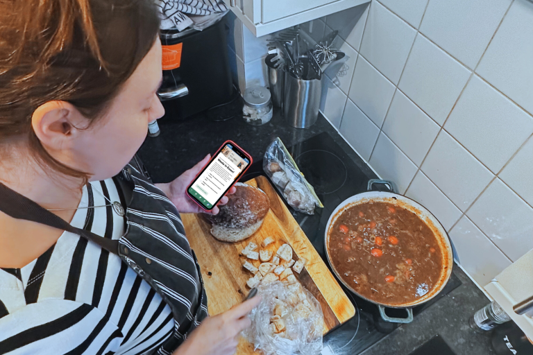 Woman wearing an apron and cooking in the kitchen, she is holding a phone in her left hand.