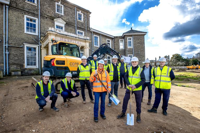 A large group of people in high-visibility jackets and hard hats holding shovels standing in front of a historic building