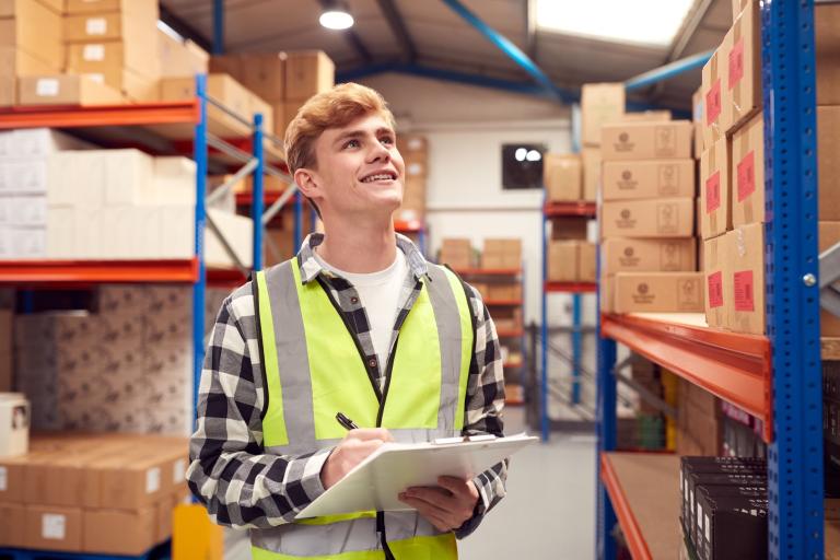 A young man wearing a high-visibility jacket standing inside a warehouse area holding a clipboard and pen.