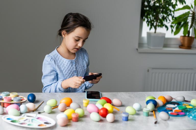 Young girl sat at a table on a phone with easter eggs and activities on the table.