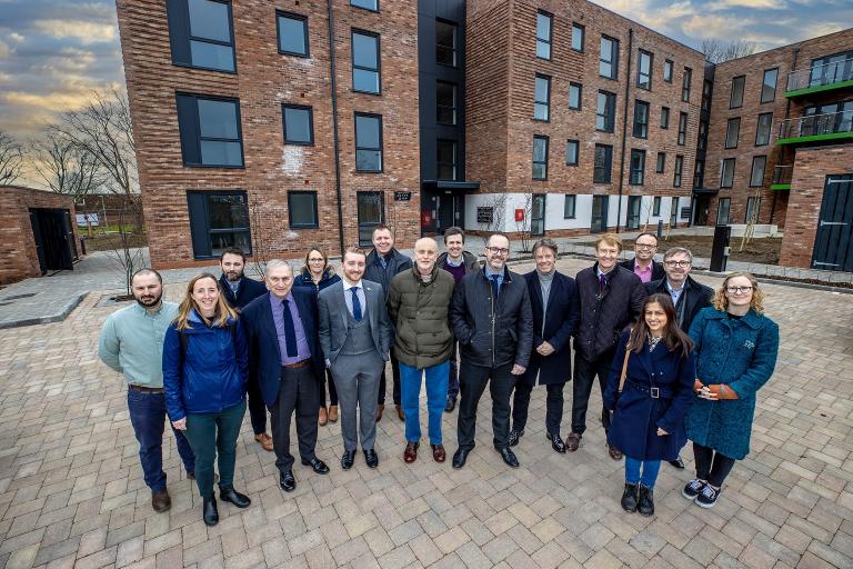 A large group of people standing in a crescent shape, in front of a collection of modern apartments.